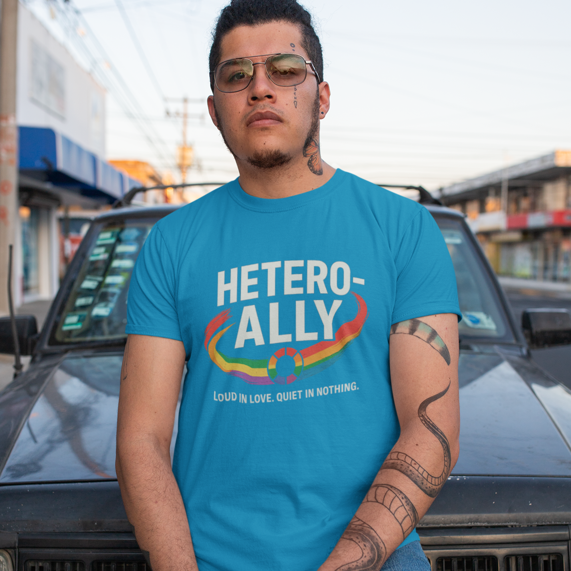A Latino man wearing a blue t-shirt, wearing a Glowfolks 'HETERO-ALLY' classic tee, chillin' in front of his vehicle.